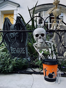 A Halloween-themed display is set in a garden with tombstones and skeletons. A realistic skull is positioned next to ironwork that spells 'Beware'. A humorous touch is added by a skeleton figure holding an orange coffee cup featuring Halloween illustrations, including a skeleton and bats. The backdrop is a classic white house with a yellow door.