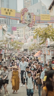 A bustling street market with numerous people walking and shopping. Various shops line the sides, displaying vibrant merchandise and signs in multiple languages. Tall modern buildings rise in the background, contrasting with the traditional street market setting. The scene is lively and crowded, with a diverse mix of pedestrians.