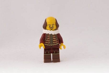A small LEGO figure with a yellow face and hands, dressed in a dark red outfit with gold patterns resembling a historical costume. It features a white ruffled collar and brown hair.