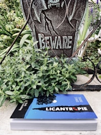 A weathered sign with the word 'BEWARE' and a menacing figure is set among green plants. Below the sign, there is a book titled 'LICANTROPIE' which features a blue cover with a silhouette of a werewolf and a full moon against a starry night sky.
