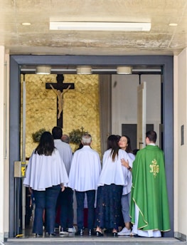 A group of people wearing white robes standing inside a church entrance with a priest in a green vestment on the right. A crucifix is mounted on a textured wall in the background. Some individuals are interacting warmly, possibly hugging.