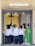 A group of people wearing white robes standing inside a church entrance with a priest in a green vestment on the right. A crucifix is mounted on a textured wall in the background. Some individuals are interacting warmly, possibly hugging.