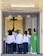 A group of people wearing white robes standing inside a church entrance with a priest in a green vestment on the right. A crucifix is mounted on a textured wall in the background. Some individuals are interacting warmly, possibly hugging.