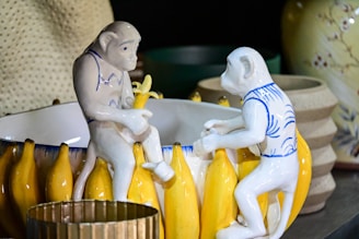 Two ceramic monkey figurines are interacting with a bowl that is decorated with banana motifs. The figurines are painted with blue and white patterns and appear to be holding bananas. The background includes a variety of ceramic and pottery objects in muted tones.