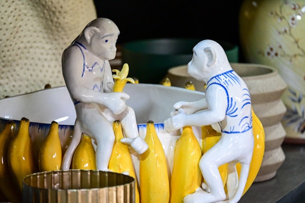 Two ceramic monkey figurines are interacting with a bowl that is decorated with banana motifs. The figurines are painted with blue and white patterns and appear to be holding bananas. The background includes a variety of ceramic and pottery objects in muted tones.