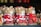 A collection of straw-crafted festive dog ornaments dressed in red and white Santa Claus outfits, with some wearing hats and others adorned with scarves. They feature button eyes and black noses, standing together on a shelf as part of holiday decorations.