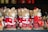 A collection of straw-crafted festive dog ornaments dressed in red and white Santa Claus outfits, with some wearing hats and others adorned with scarves. They feature button eyes and black noses, standing together on a shelf as part of holiday decorations.