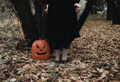 The scene features a carved pumpkin with a sinister face placed on the ground among fallen autumn leaves. A person in a long black dress standing next to the pumpkin, with only their lower legs visible, adds a mysterious element to the woodland setting.