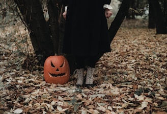 The scene features a carved pumpkin with a sinister face placed on the ground among fallen autumn leaves. A person in a long black dress standing next to the pumpkin, with only their lower legs visible, adds a mysterious element to the woodland setting.