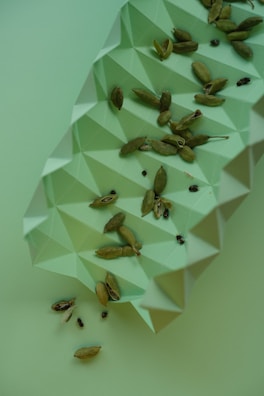 Several cardamom pods are scattered on a light green, geometric, textured surface. The surface has a three-dimensional origami-like pattern, creating an interesting visual contrast with the organic shape of the cardamom pods.