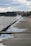 A concrete walkway runs alongside a body of water with waves crashing against it. The walkway is wet and lined with a series of metal posts. In the distance, there are trees and buildings under a cloudy sky, suggesting an urban waterfront area.