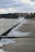 A concrete walkway runs alongside a body of water with waves crashing against it. The walkway is wet and lined with a series of metal posts. In the distance, there are trees and buildings under a cloudy sky, suggesting an urban waterfront area.