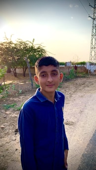 A young person stands on a dirt path with a serene expression, wearing a blue shirt. The background features lush greenery, trees, and a clear sky, with gentle sunlight creating a warm atmosphere. Power lines are visible to the right, adding an element of rural infrastructure.