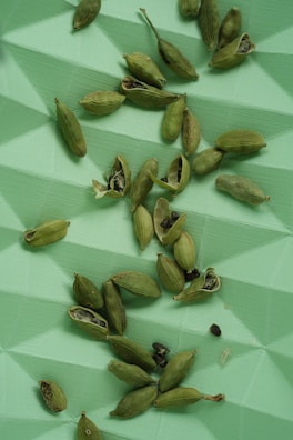 Green cardamom pods scattered across a textured, geometric green surface. Some pods are whole, while others are open, revealing small black seeds.