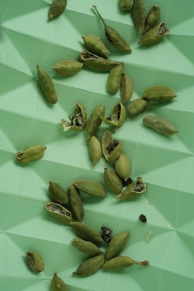Green cardamom pods scattered across a textured, geometric green surface. Some pods are whole, while others are open, revealing small black seeds.