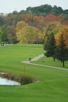 A lush green golf course is surrounded by a variety of trees displaying vibrant autumn colors. A small pavilion stands amidst the trees. A path winds through the course where a golf cart and a figure carrying a golf bag are seen.