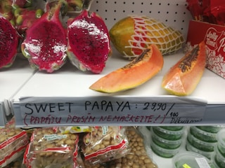 Several exotic fruits are displayed on a supermarket shelf. There are cut pieces of dragon fruits with dark seeds and vibrant pink flesh, as well as slices of papaya. A handwritten sign notes 'Sweet Papaya: 29,90' alongside a request not to squeeze the papayas. Below the fruits, packages of brown mushrooms are visible, and a box of Jolly Time popcorn is partly seen on the side.