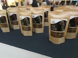 Several brown paper bags with clear windows displaying dried worms are arranged on a dark surface. The bags are labeled with text and graphics, likely indicating the product and brand name. In the background, blurred figures and a bright interior are visible, suggesting a market or exhibition setting.