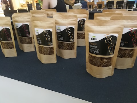 Several brown paper bags with clear windows displaying dried worms are arranged on a dark surface. The bags are labeled with text and graphics, likely indicating the product and brand name. In the background, blurred figures and a bright interior are visible, suggesting a market or exhibition setting.