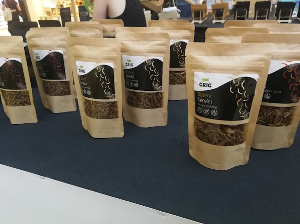 Several brown paper bags with clear windows displaying dried worms are arranged on a dark surface. The bags are labeled with text and graphics, likely indicating the product and brand name. In the background, blurred figures and a bright interior are visible, suggesting a market or exhibition setting.