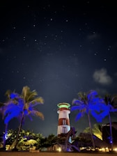 A lighthouse is surrounded by palm trees under a starry night sky. The lighthouse is illuminated with bright lights, featuring red and white stripes. The palm trees are lit with blue and green hues, creating a vibrant contrast against the dark sky.
