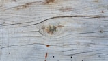Weathered wooden surface with visible grain patterns and a prominent knot in the center. The wood appears aged, with natural cracks and some reddish-brown discolorations.