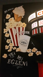 A decorative wall depicts a whimsical design featuring a large striped popcorn bucket overflowing with popcorn, alongside a tall ice cream cone. A cup with a logo and surrounding text is part of the composition. A retro cinema sign with lights displays several words.