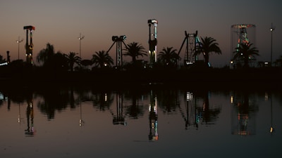 An amusement park is silhouetted against a darkening sky at dusk, with various rides and palm trees reflecting in a calm body of water. The rides are subtly illuminated with colorful lights, adding a contrast to the twilight tones.