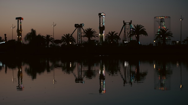 An amusement park is silhouetted against a darkening sky at dusk, with various rides and palm trees reflecting in a calm body of water. The rides are subtly illuminated with colorful lights, adding a contrast to the twilight tones.
