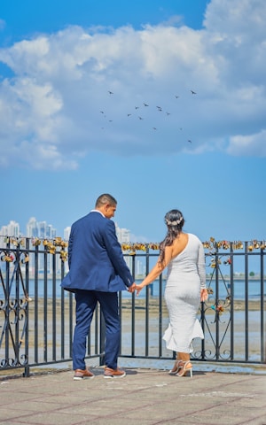 A couple standing hand in hand on a waterfront promenade, with love locks attached to a railing. They are dressed formally, the man in a blue suit and the woman in a white dress. The background features a cloudy blue sky and distant city buildings. Birds are flying above in the sky.