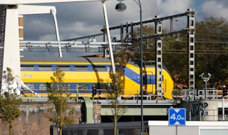 A yellow and blue double-decker train passes over a bridge amidst railway infrastructure. Overhead lines and signals are visible, along with some structural elements of the bridge. Trees and a graffitied wall are in the foreground, and a lamppost is positioned near the train. A blue road sign indicating a height limit of 4.0 meters is present.