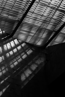 Shadow patterns are cast on a textured wall by an overhead structure with slatted elements, creating an interplay of light and shadows in a geometric pattern.
