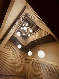 An architectural interior featuring a geometric, wooden ceiling design with multiple spherical white lights hanging down. The structure creates a sense of depth and symmetry with vertical and diagonal slatted wood panels.