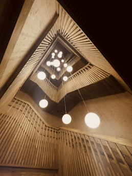 An architectural interior featuring a geometric, wooden ceiling design with multiple spherical white lights hanging down. The structure creates a sense of depth and symmetry with vertical and diagonal slatted wood panels.