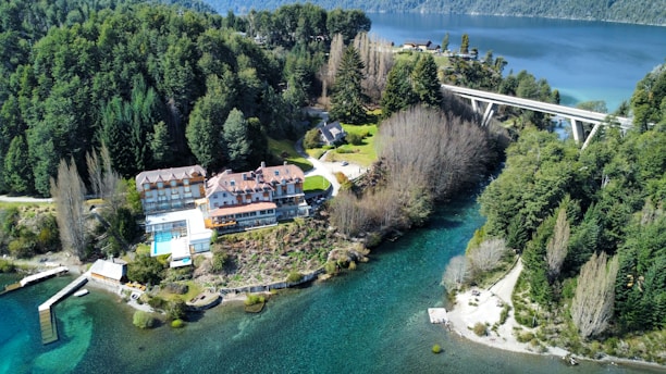 An aerial view of a large building complex surrounded by lush green forests and a serene body of water. A bridge crosses over the water, connecting two forested areas. The building features multiple sections, including a pool area and a dock extending into the clear blue water.