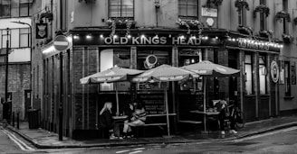 A black and white image featuring a corner pub with the name 'Old Kings Head'. The establishment has a traditional facade with illuminated signage, hanging plants, and outdoor seating under umbrellas. Two people are seated at a table, one looking at their phone. A bicycle is parked nearby. The street sign 'Scrutton St.' is visible, and the wet pavement suggests recent rain.