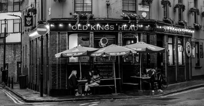 A black and white image featuring a corner pub with the name 'Old Kings Head'. The establishment has a traditional facade with illuminated signage, hanging plants, and outdoor seating under umbrellas. Two people are seated at a table, one looking at their phone. A bicycle is parked nearby. The street sign 'Scrutton St.' is visible, and the wet pavement suggests recent rain.