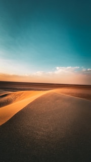 A vast desert landscape with gently sloping sand dunes highlighted by warm sunlight. The sky above is expansive, transitioning from a deep blue to a soft orange near the horizon, dotted with a few wispy clouds.