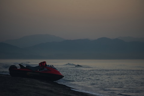 A red jet ski is parked on a dark sandy beach near a calm ocean. The scene features a backdrop of misty mountains under a dusky sky, suggesting a serene and tranquil setting.