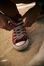 A person is tying the shoelaces of a red canvas sneaker with white rubber sole. The sneaker has a small decorative item resembling a donut attached to the laces.