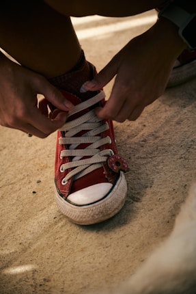 A person is tying the shoelaces of a red canvas sneaker with white rubber sole. The sneaker has a small decorative item resembling a donut attached to the laces.