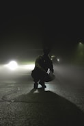 A silhouette of a person crouching on a deserted road with a car's headlights creating a dramatic backlight through thick fog. The scene is set at night, emphasizing a mysterious and tense atmosphere.