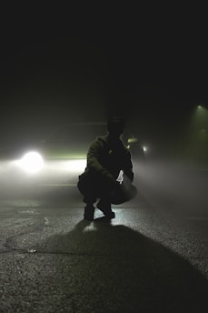 A silhouette of a person crouching on a deserted road with a car's headlights creating a dramatic backlight through thick fog. The scene is set at night, emphasizing a mysterious and tense atmosphere.