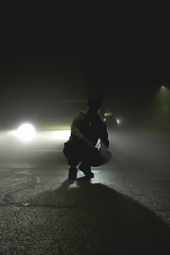 A silhouette of a person crouching on a deserted road with a car's headlights creating a dramatic backlight through thick fog. The scene is set at night, emphasizing a mysterious and tense atmosphere.