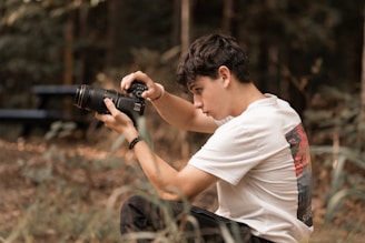 A young person with dark hair is crouching and intently focused on capturing a photograph with a DSLR camera in a natural, outdoor setting with blurred foliage in the background.