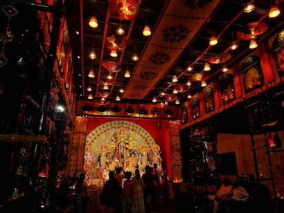 An elaborately decorated indoor setting features a large and ornate statue of a deity with multiple arms, surrounded by smaller statues. The decor includes intricate patterns, vibrant colors, and traditional motifs on the walls and ceiling, all illuminated by warm, ambient lighting. A crowd of people observes the scene, adding to a festive atmosphere.