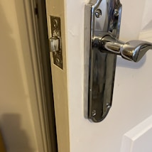 A close-up of a door with a shiny metal handle and latch, slightly ajar. The door is white, and the metal latch is installed on the frame, which appears worn around the latch slot.