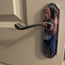 A close-up of a polished chrome door handle attached to a light-colored door. The handle has a slightly curved design and is affixed to a decorative base plate with screws. Reflections on the handle suggest a mix of colors, possibly from the surrounding environment.