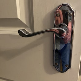 A close-up of a polished chrome door handle attached to a light-colored door. The handle has a slightly curved design and is affixed to a decorative base plate with screws. Reflections on the handle suggest a mix of colors, possibly from the surrounding environment.