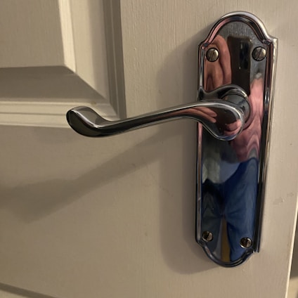 A close-up of a polished chrome door handle attached to a light-colored door. The handle has a slightly curved design and is affixed to a decorative base plate with screws. Reflections on the handle suggest a mix of colors, possibly from the surrounding environment.